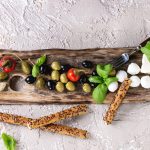 Mediterranean appetizer antipasti board with green black olives, feta cheese, mozzarella, capers, pepper, basil with grissini bread sticks over beige concrete texture background. Top view with space