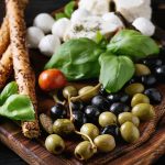Mediterranean appetizer antipasti board with green black olives, feta cheese, mozzarella, capers, pepper, basil with grissini bread sticks over black wood burnt background. Close up